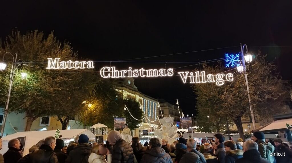 I mercatini di Natale a Matera con la folla che si aggira in Piazza Vittorio Veneto - scritta luminosa Matera Christmas Village sovrasta la scena