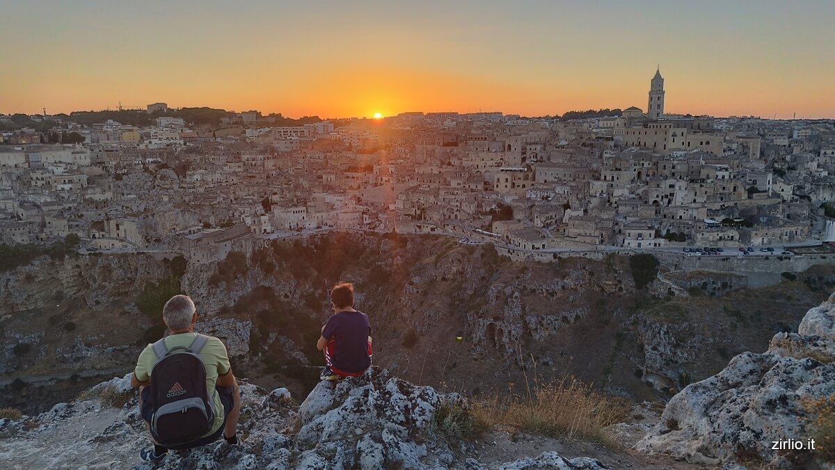 La città di Matera al tramonto con il sole a metà mentre scompare dietro di essa. Due viaggiatori di età molto diversa la ammirano estasiati dal belvedere. Tra loro e la città c'è il vuoto del canyon della Gravina.